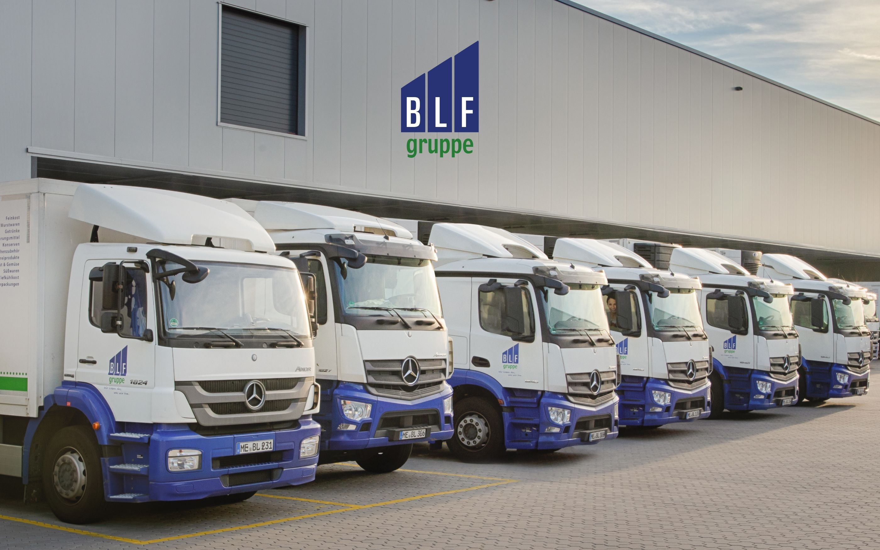 Six white trucks with blue details are parked side by side in front of a modern industrial building. The BLF Group logo in blue and green is visible on the facade.