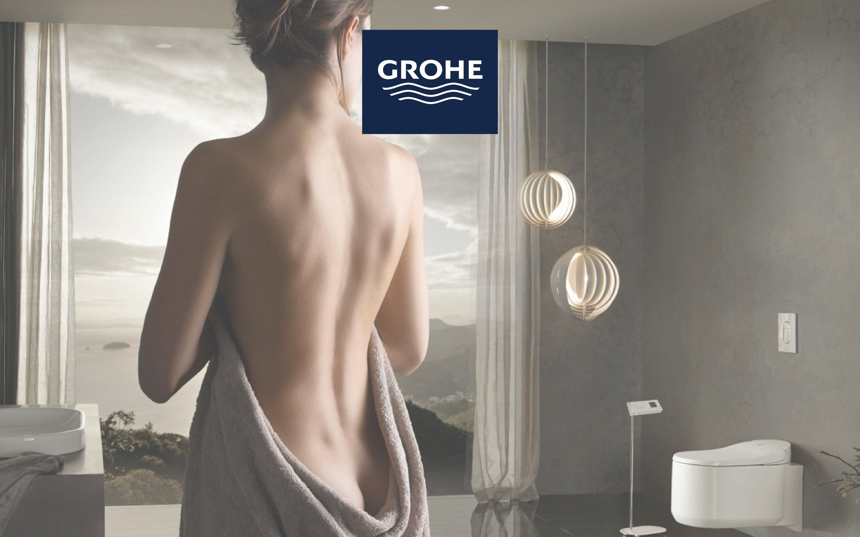 Modernes Badezimmer mit großem Fenster und Blick auf eine Landschaft. Im Vordergrund eine Person mit Handtuch um die Hüfte, im Hintergrund Designer-Waschtisch, wandhängendes WC und zwei runde Pendelleuchten. Oben das Grohe-Logo.