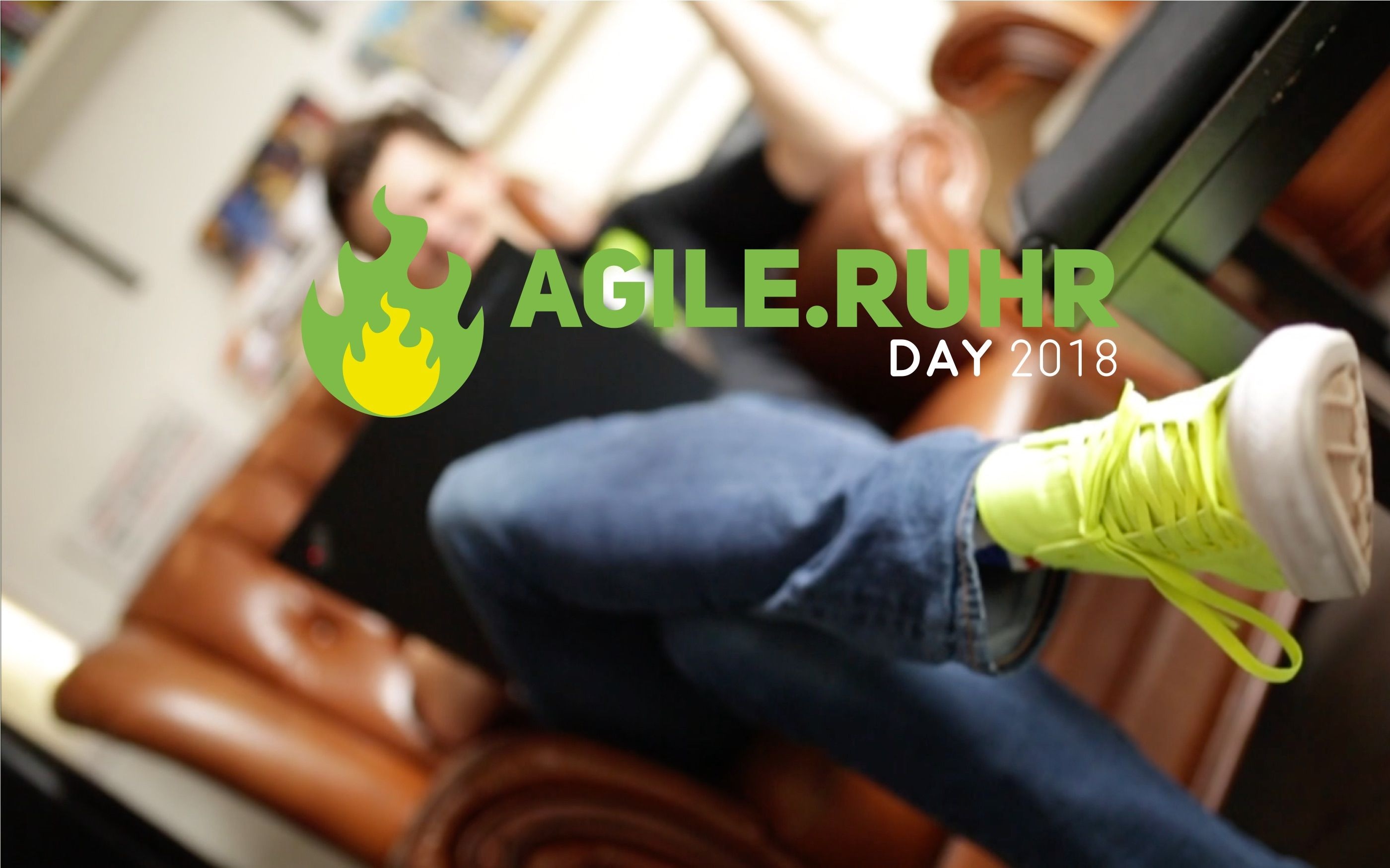Verschwommenes Foto einer Person, die lässig auf einem Sofa sitzt. Davor das Logo des Agile.Ruhr Day 2018