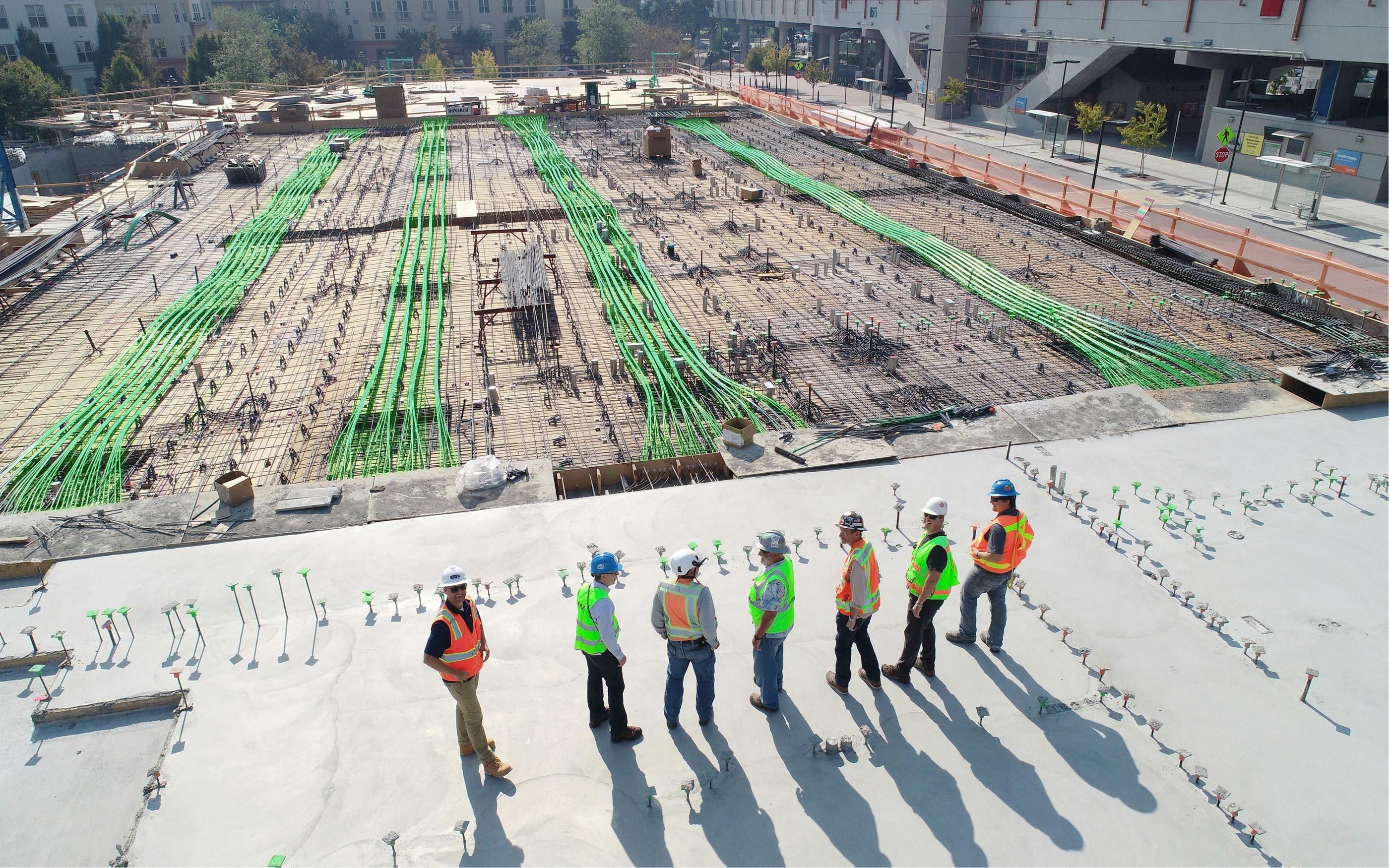 Six people wearing safety vests and protective helmets are standing together on a construction site, talking to each other. In the background, large concrete surfaces, numerous green installation pipes, and construction materials can be seen.