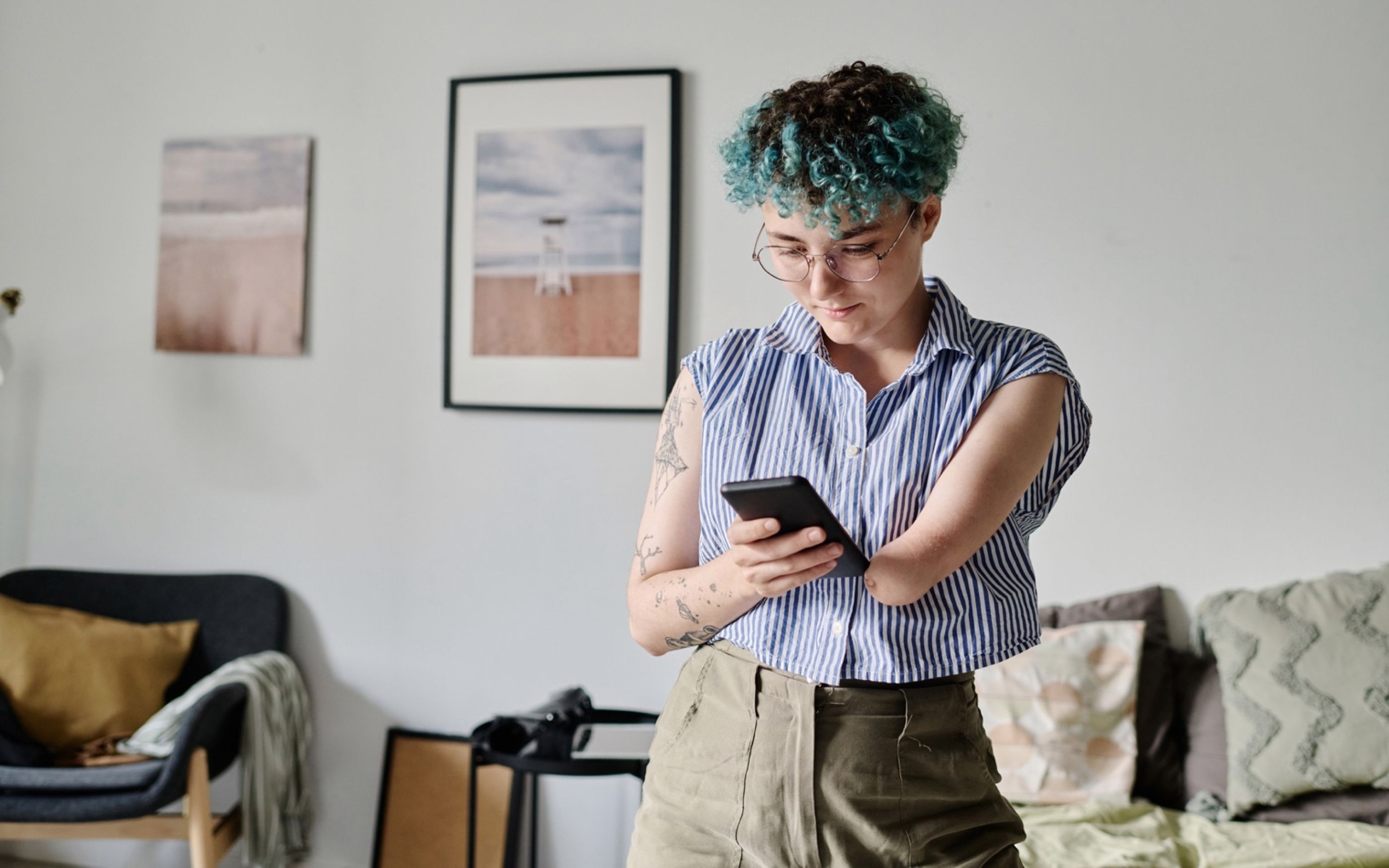A woman with short, dyed blue hair stands in a living room, looking at her cell phone, which she holds in her right hand. Her left arm, amputated from the elbow, is supporting the phone.