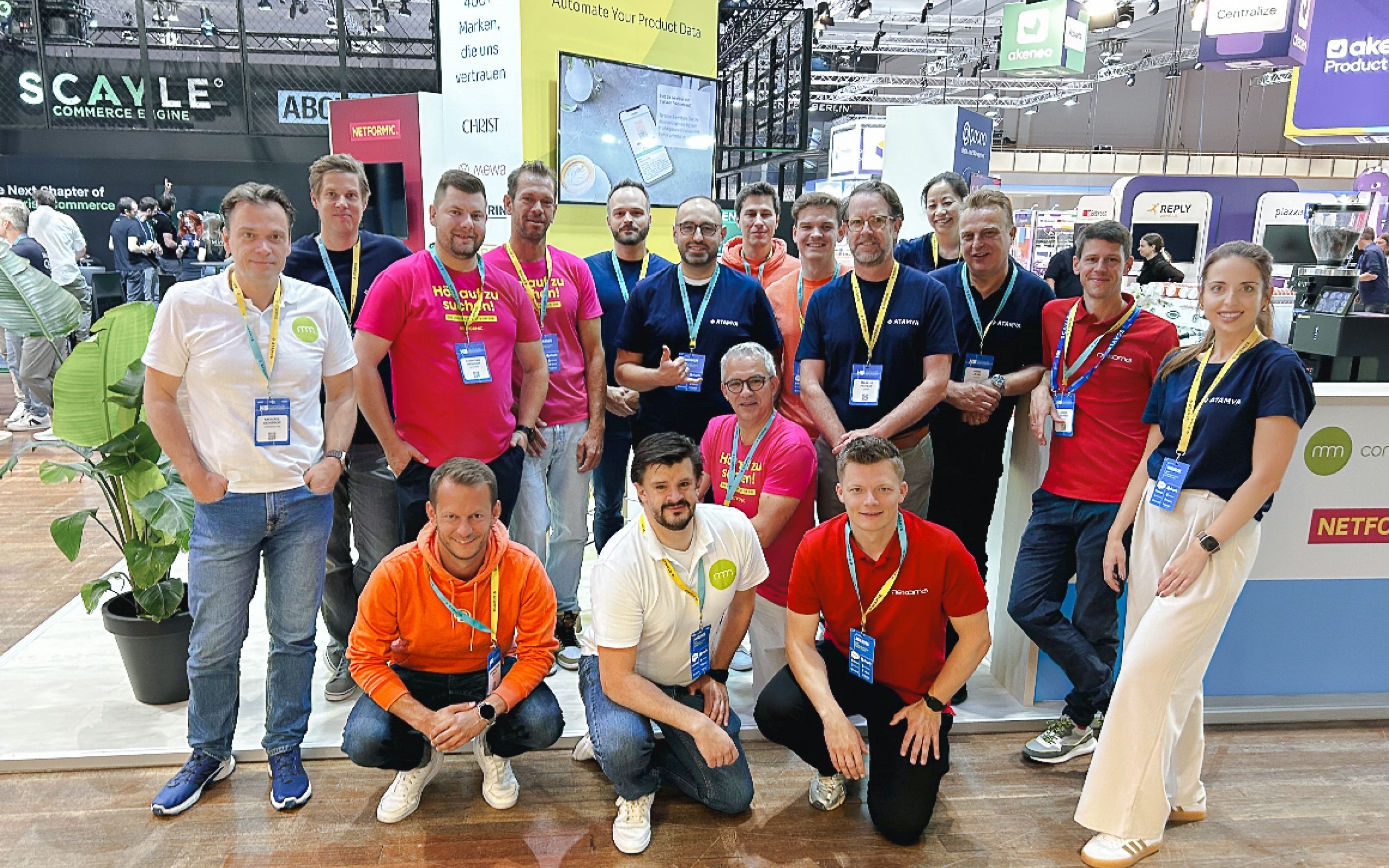 Group photo of trade fair team in front of a booth with banners and displays; people are wearing colored T-shirts with company logos and name tags, with other booths and visitors in the background.