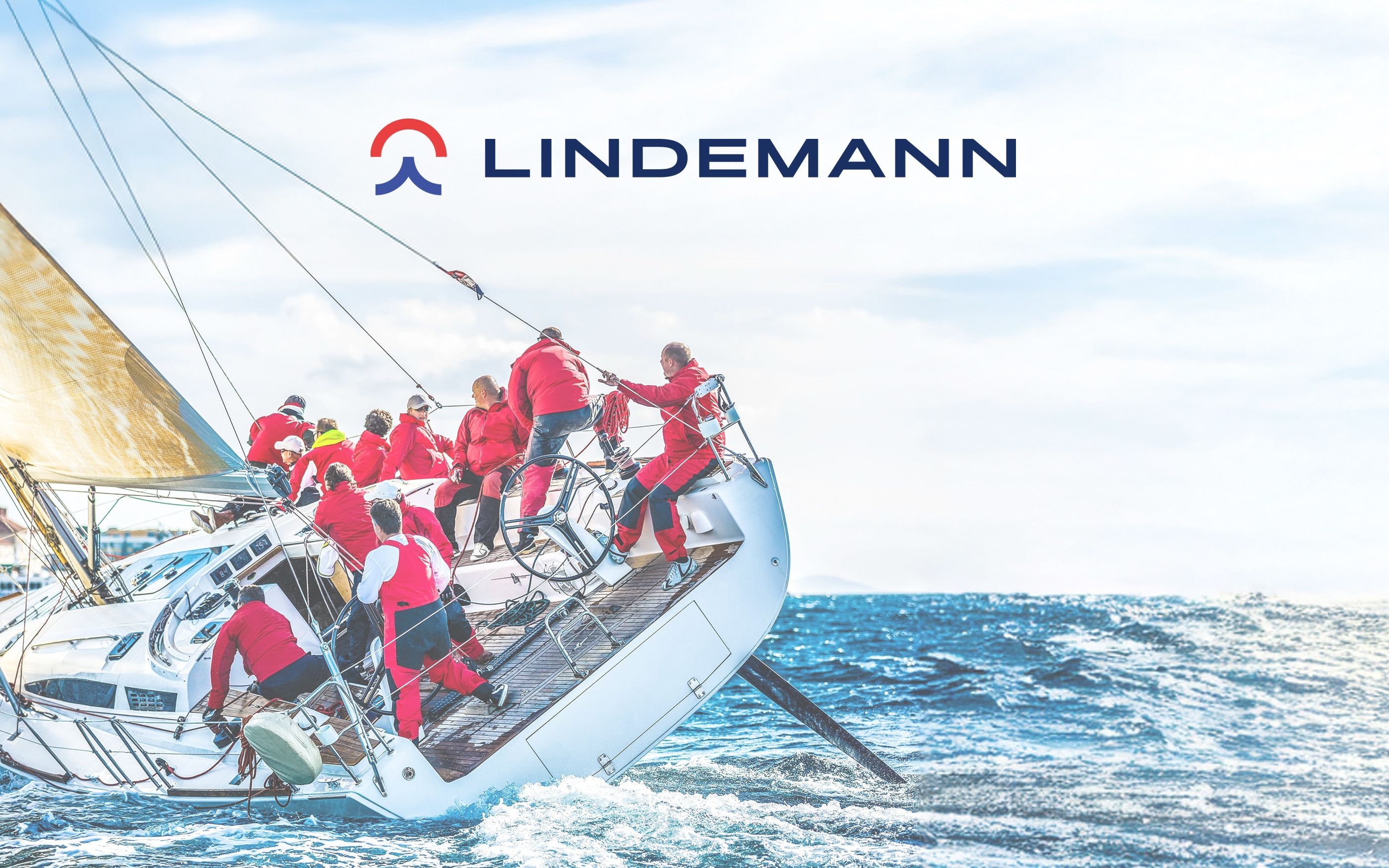 Sailboat with crew in red jackets on stormy sea, leaning heavily to starboard. Blue sky with clouds in the background. Lindemann logo in blue and red at the top.