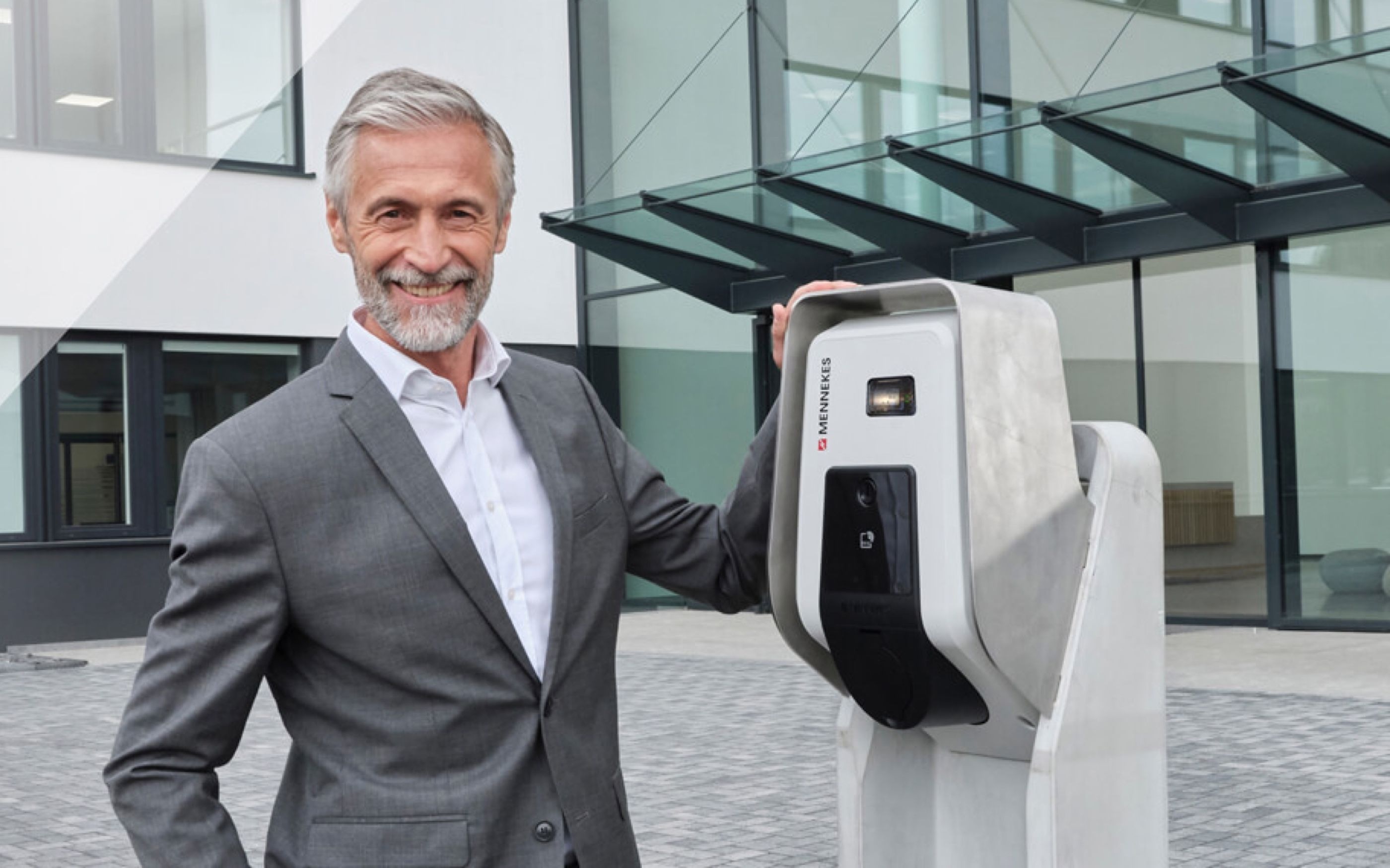 Person in grauem Anzug steht vor einem modernen Gebäude und legt die Hand auf eine graue Ladesäule von Mennekes mit integriertem Display und Anschluss für Elektrofahrzeuge.