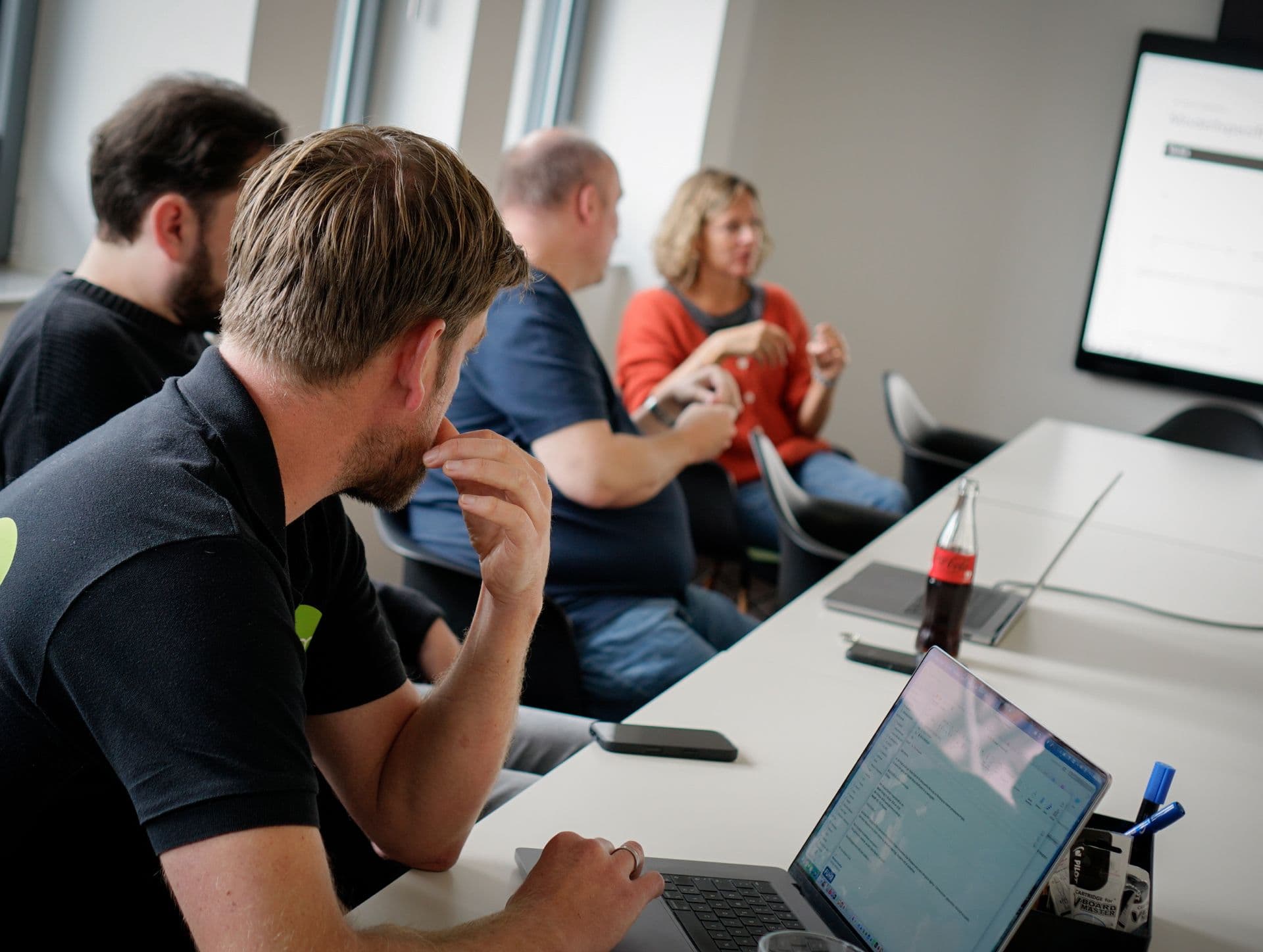 People are seated around a conference table in a meeting room, discussing in front of a display screen. Laptops and work materials are placed on the table.