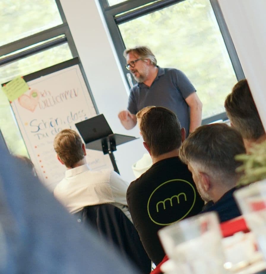 Person hält einen Vortrag vor einer kleinen Gruppe von Teilnehmenden in einem Workshop- oder Networking-Format. Im Hintergrund sind eine Präsentationsfläche und ein Flipchart zu sehen.