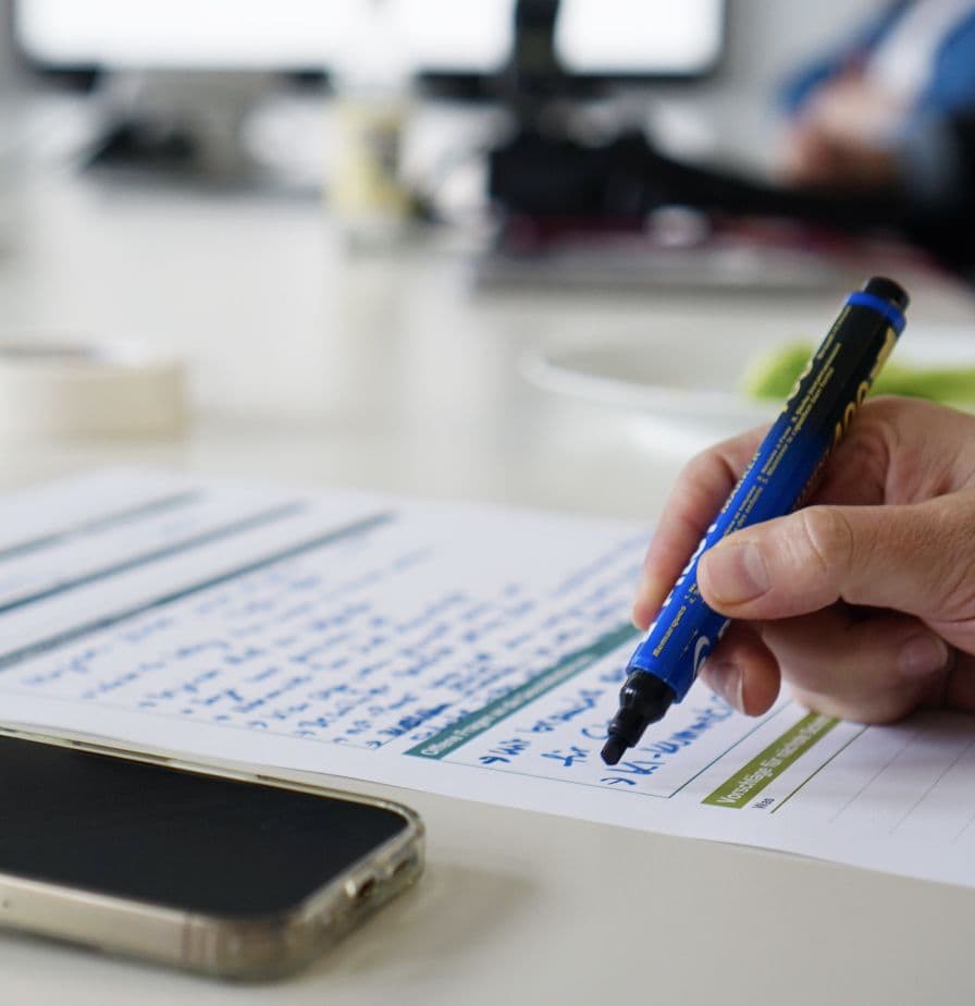 Close-up of a hand writing notes on a workshop worksheet with a marker. A smartphone is lying on the table in the foreground.