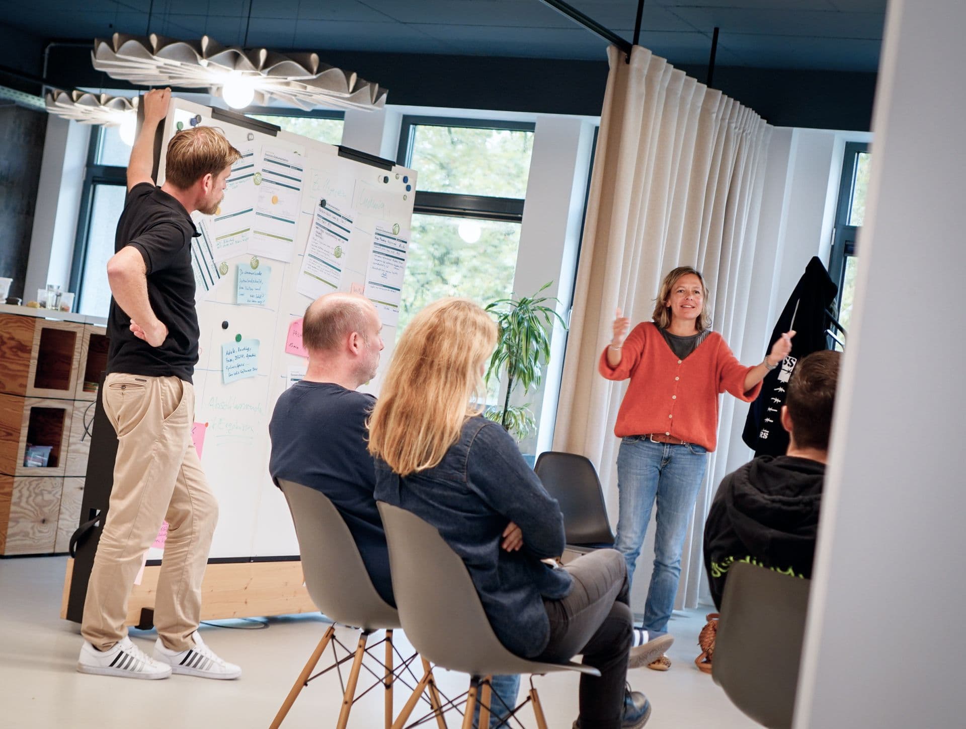 Workshop-Szene mit mehreren Teilnehmenden in einem Besprechungsraum. Eine Person präsentiert vor einer Gruppe, während auf einem Whiteboard Notizen und Arbeitsergebnisse sichtbar sind.