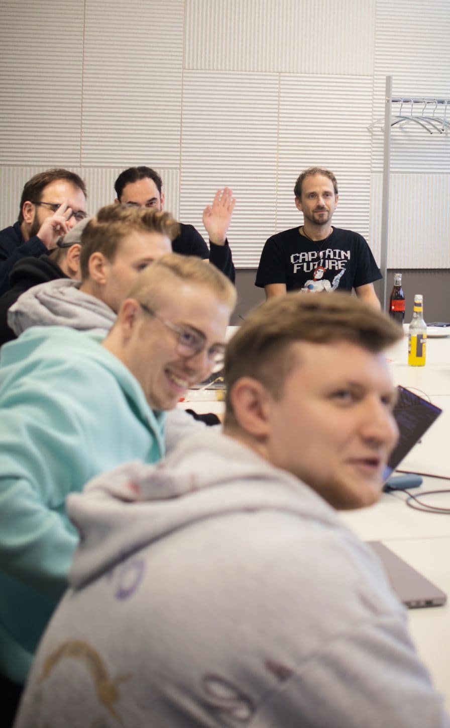 Gruppe von Teilnehmenden sitzt an einem Konferenztisch während eines Workshops oder Teammeetings. Mehrere Personen hören zu, arbeiten mit Laptops oder beteiligen sich aktiv an der Diskussion.