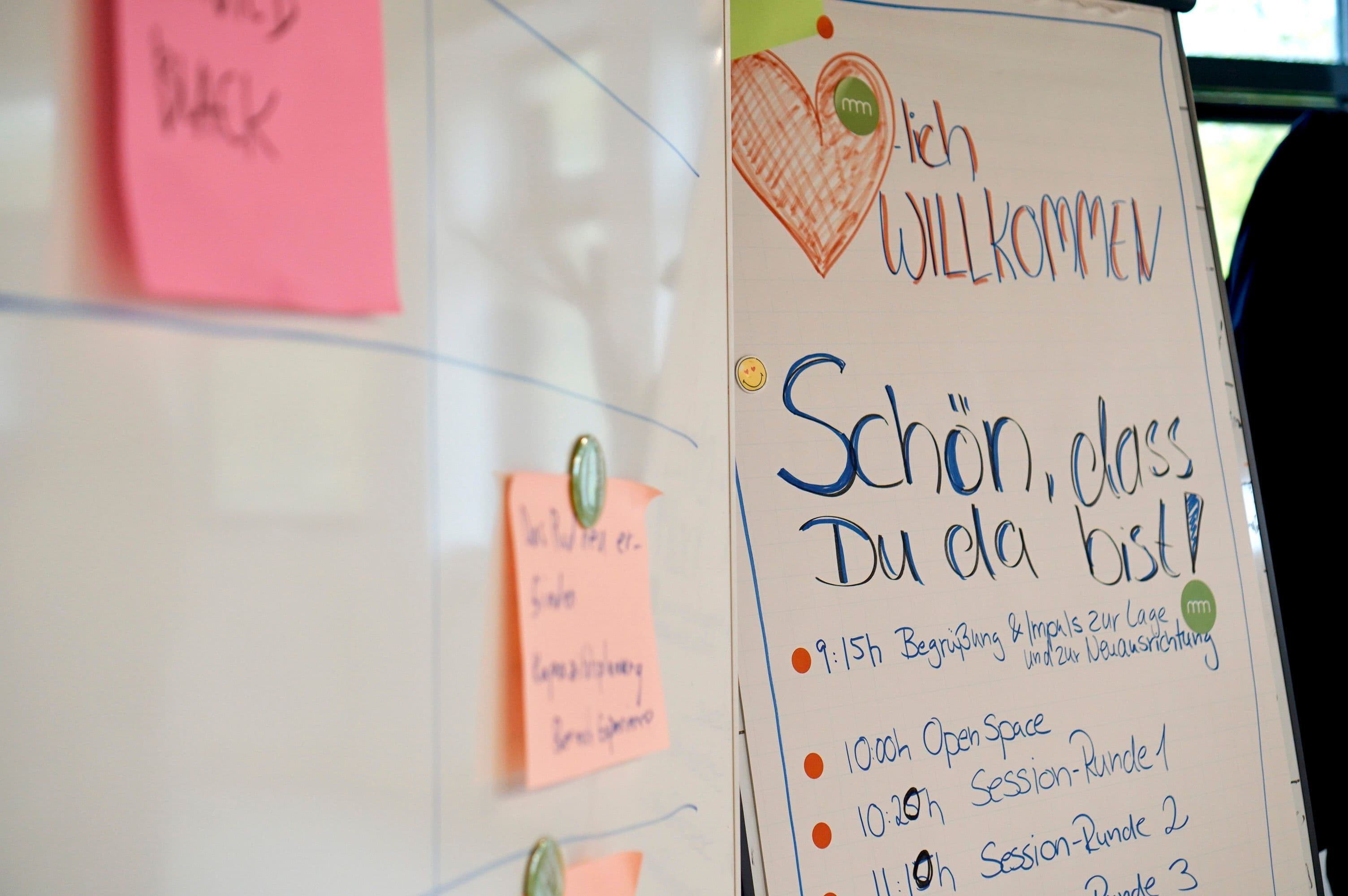 Flipchart mit Workshop-Agenda und Willkommensbotschaft neben einem Whiteboard mit Haftnotizen für die gemeinsame Arbeitsplanung.