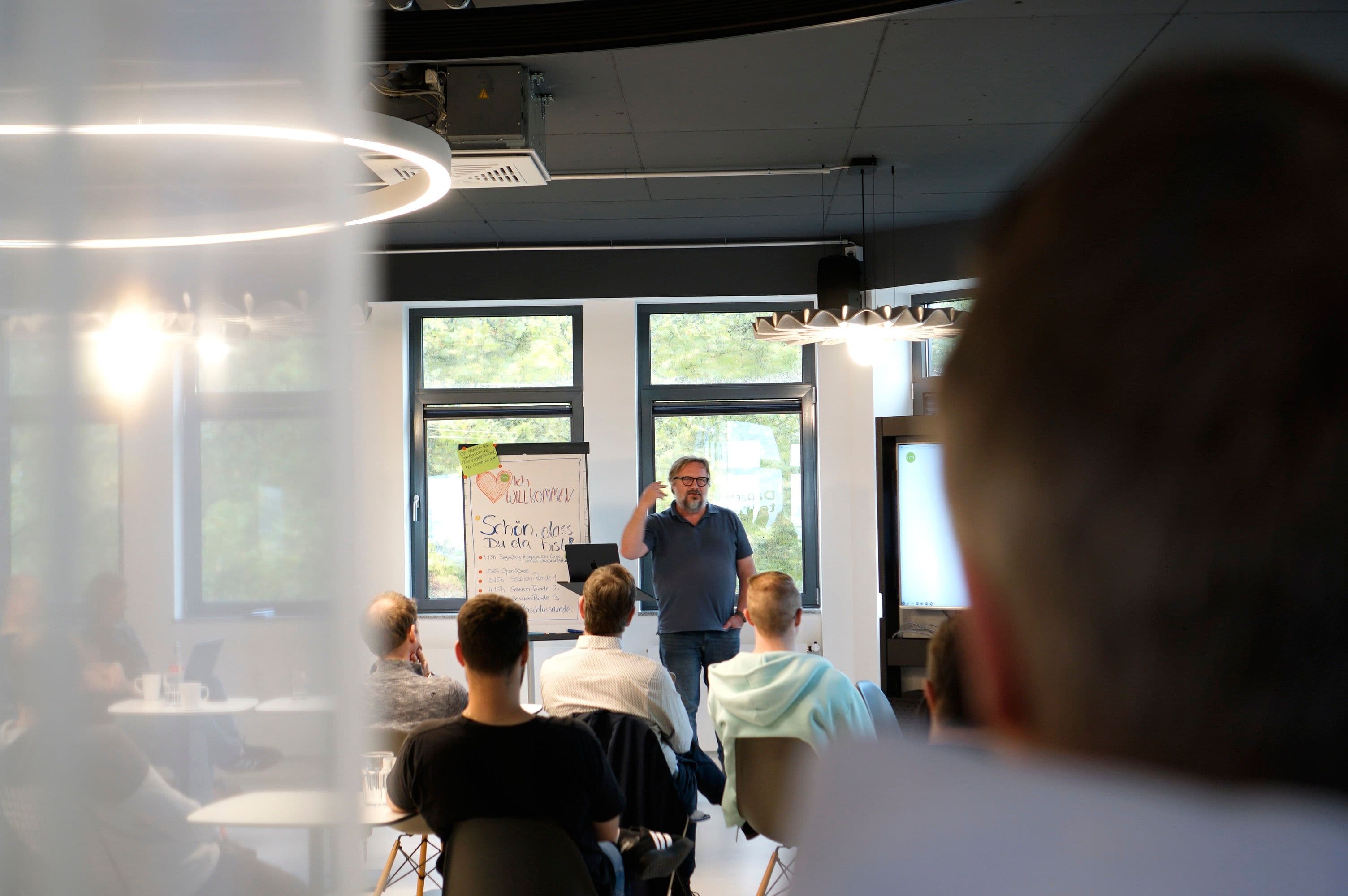 Vortragende Person spricht vor einem sitzenden Publikum während eines Workshops oder Seminars in einem modernen Veranstaltungsraum.