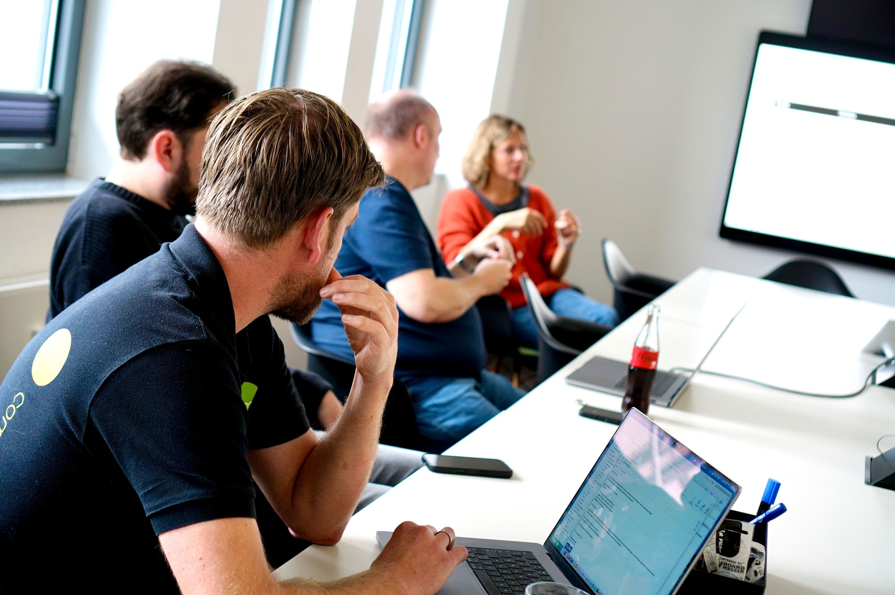Teilnehmende arbeiten mit Laptops an einem Konferenztisch und diskutieren Inhalte vor einem großen Bildschirm in einem Besprechungsraum.