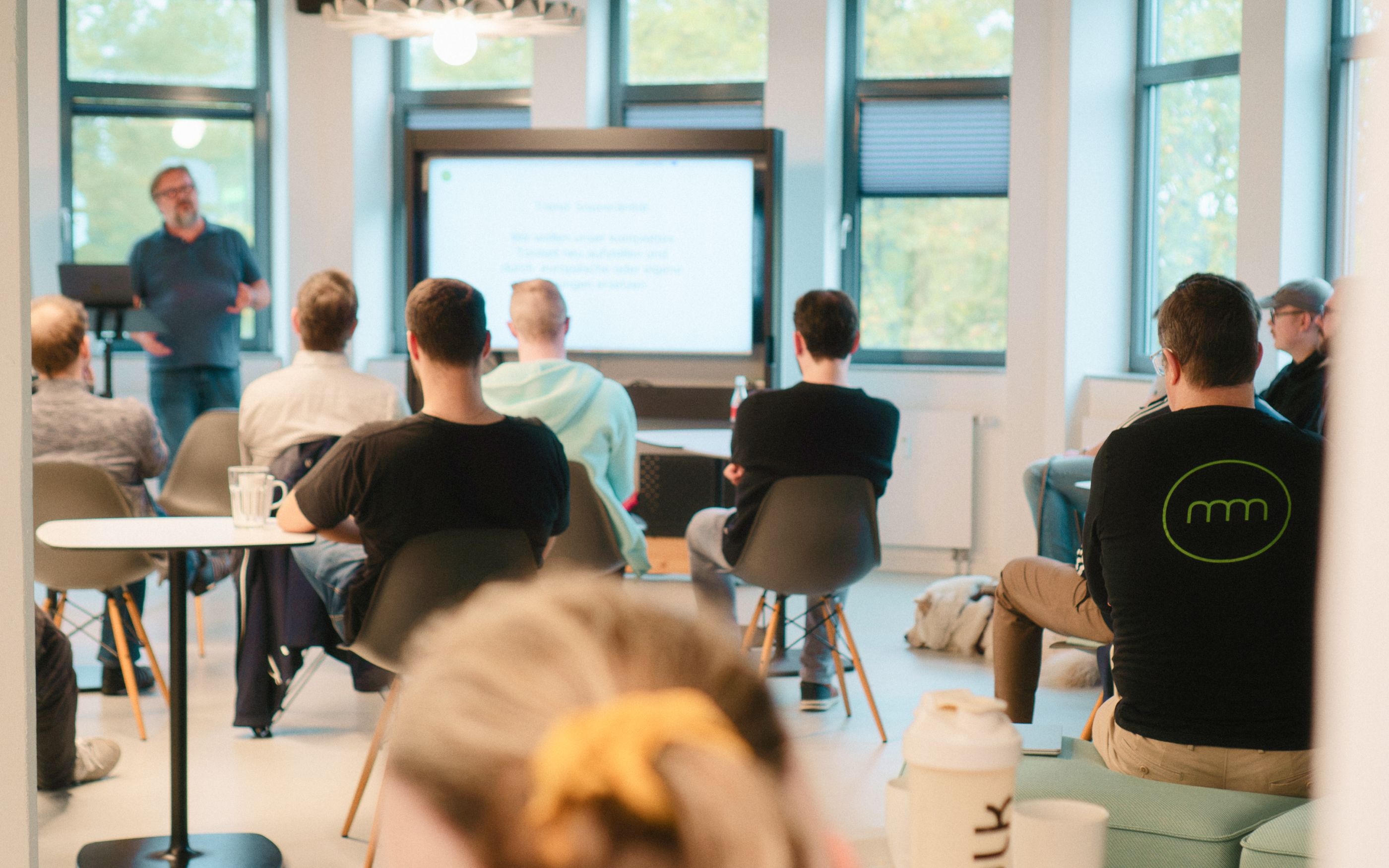 Attendees sit in a bright meeting room, listening to a presentation displayed on a large screen.