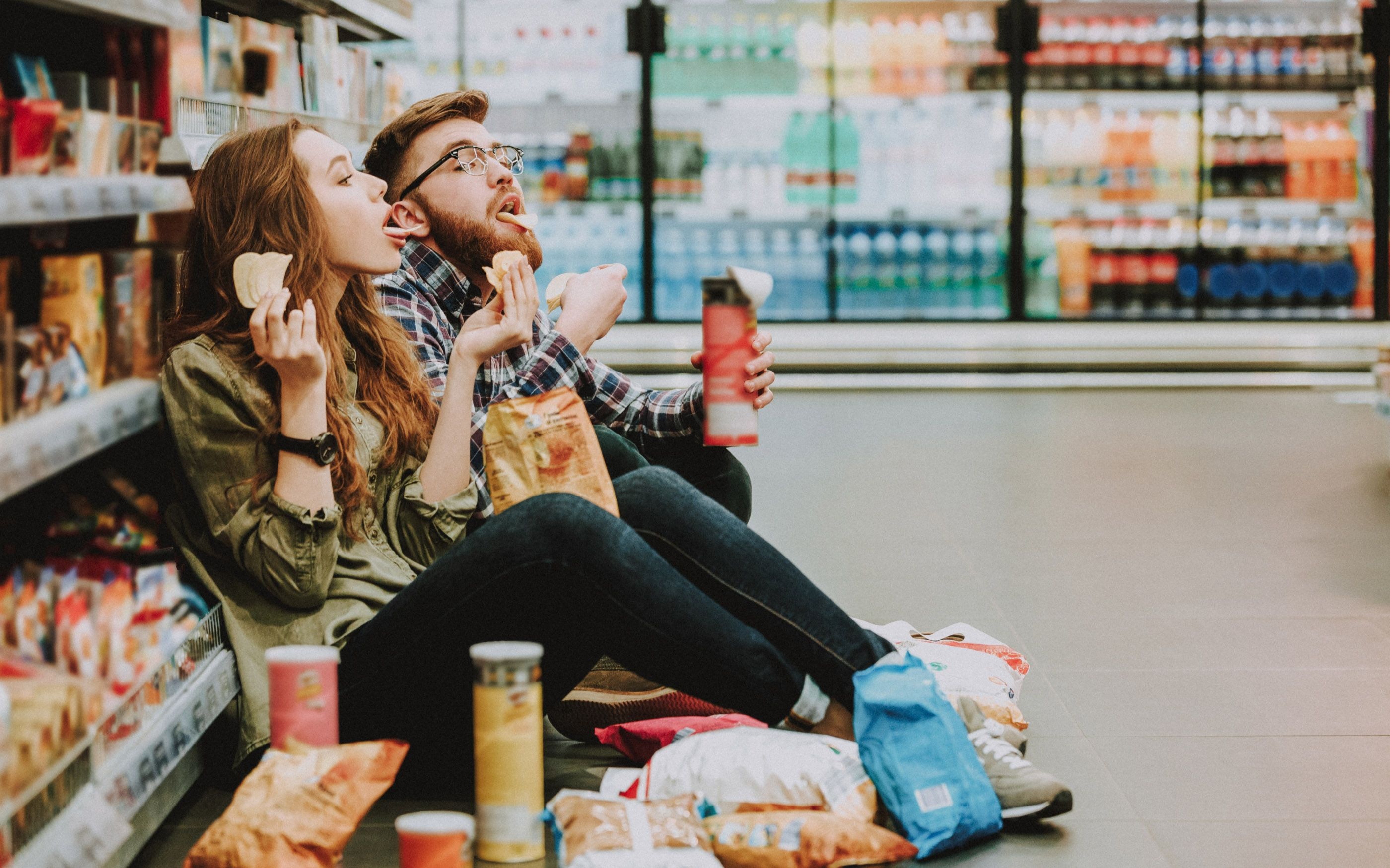 Zwei Personen sitzen auf dem Boden eines Supermarkts zwischen Regalen mit Snacks und Getränken; vor ihnen liegen geöffnete Chipstüten und weitere Snacks, beide halten Lebensmittel und Getränke in der Hand.