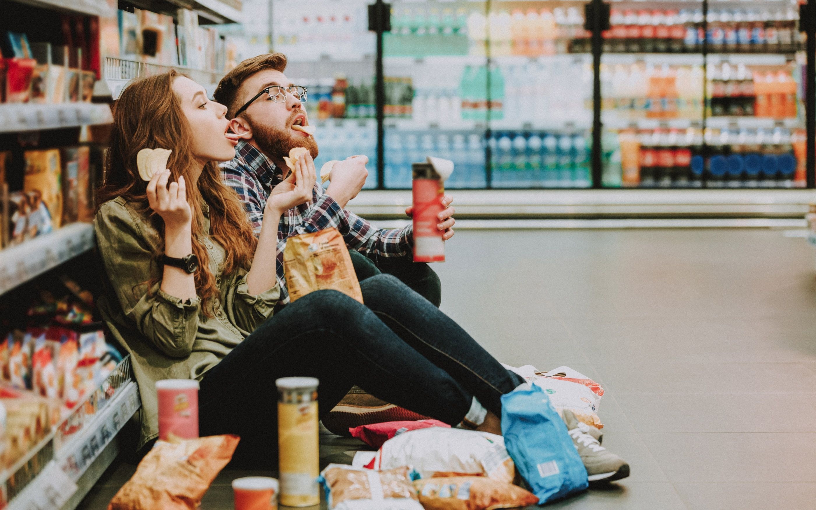 Two people are sitting on the floor of a supermarket between shelves of snacks and drinks; in front of them are open bags of chips and other snacks, and both are holding food and drinks in their hands.