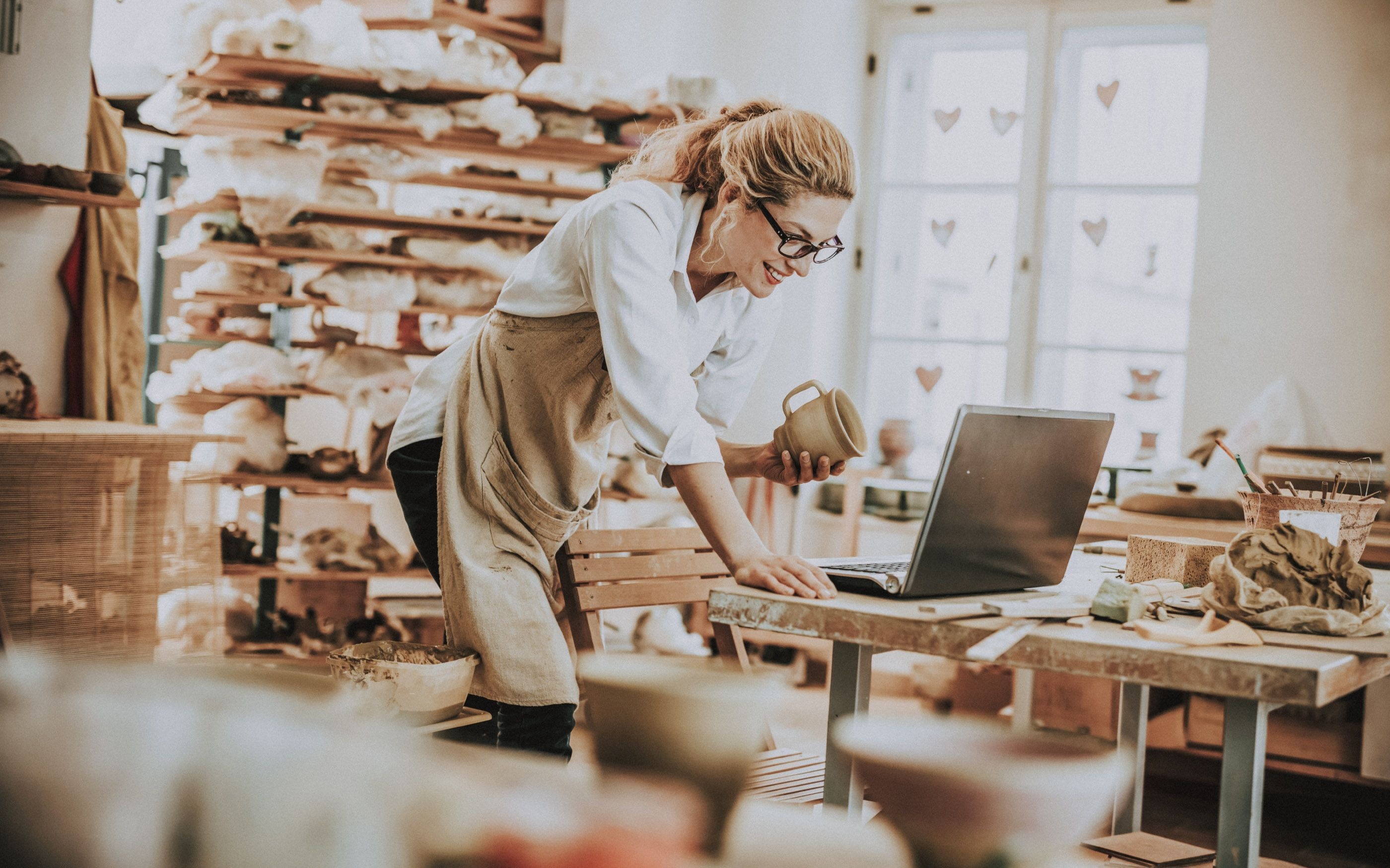 Photo of a woman standing in front of a laptop in her pottery studio with a pottery mug.