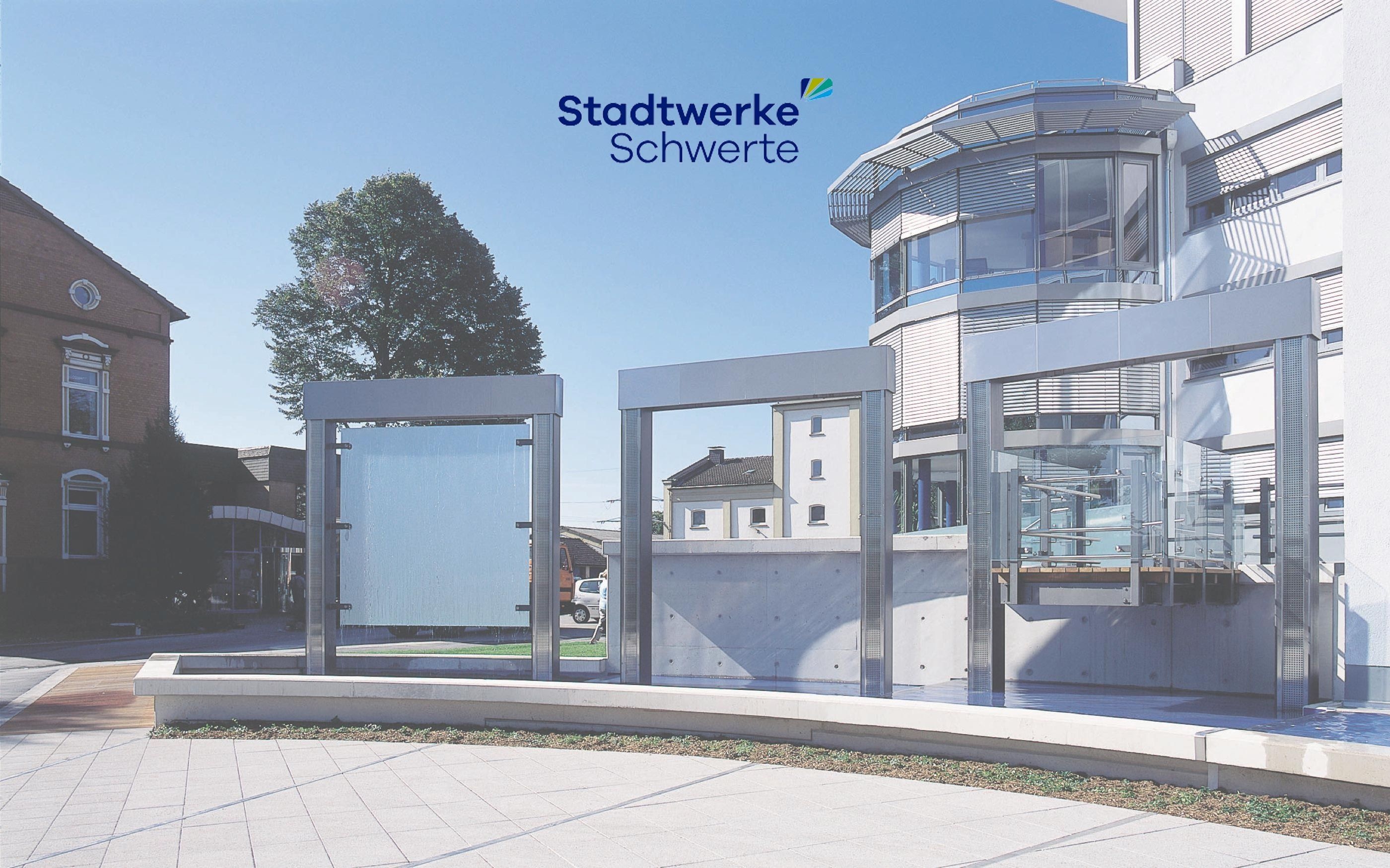 Modernes Gebäude der Stadtwerke Schwerte mit Glas- und Metallfassade, davor drei große Glasrahmen im Außenbereich. Links ein älteres Backsteingebäude, im Hintergrund Bäume und blauer Himmel. Oben das Logo ‚Stadtwerke Schwerte‘.