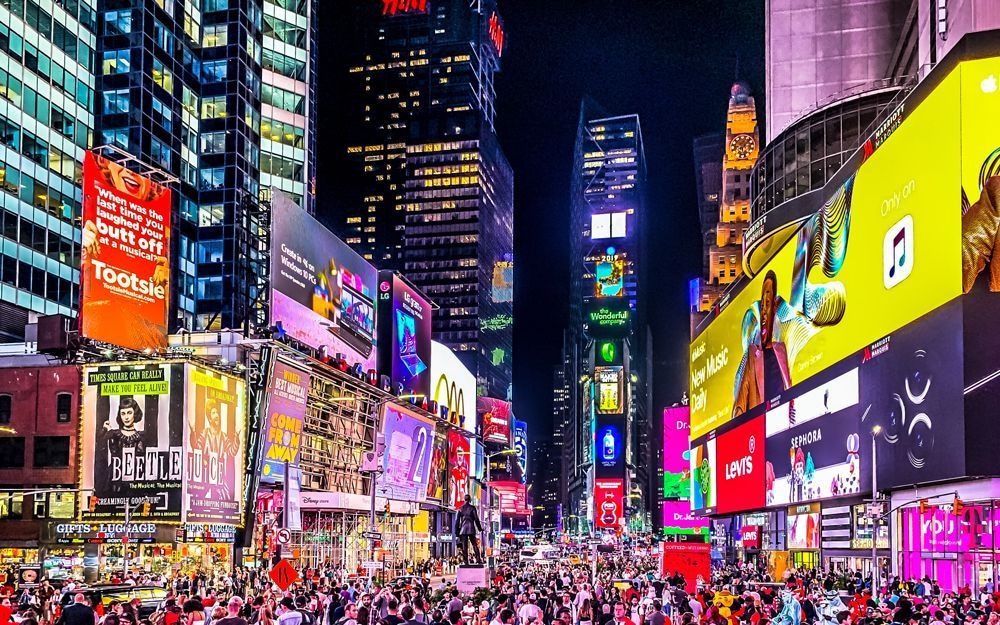 Belebte Straßenszene am Times Square in New York bei Nacht, mit zahlreichen hellen, farbenfrohen Werbetafeln und digitalen Anzeigen an Hochhäusern; viele Menschen sind auf der Straße unterwegs.